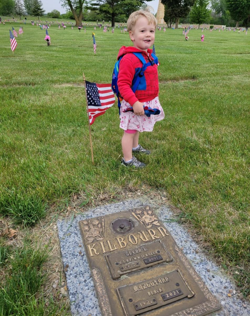 Child wearing Spiderman jacket standing by headstone with American flag