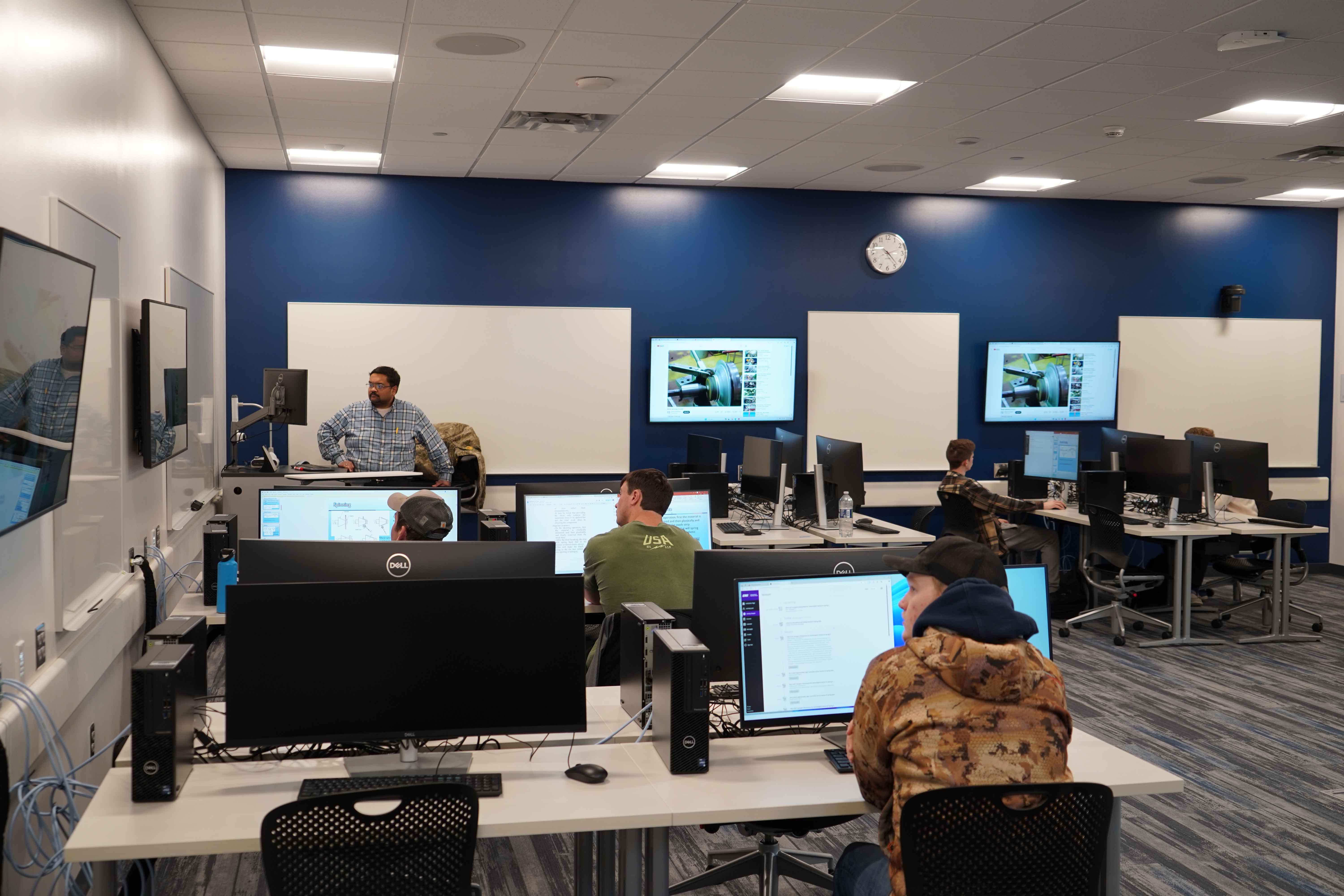 A classroom with multiple students seated at computer workstations while an instructor stands at the front near a podium and monitors. Several large screens on the walls display technical content, and students are working individually or looking toward the instructor. The room has blue accent walls, bright overhead lighting, and rows of desktop computers