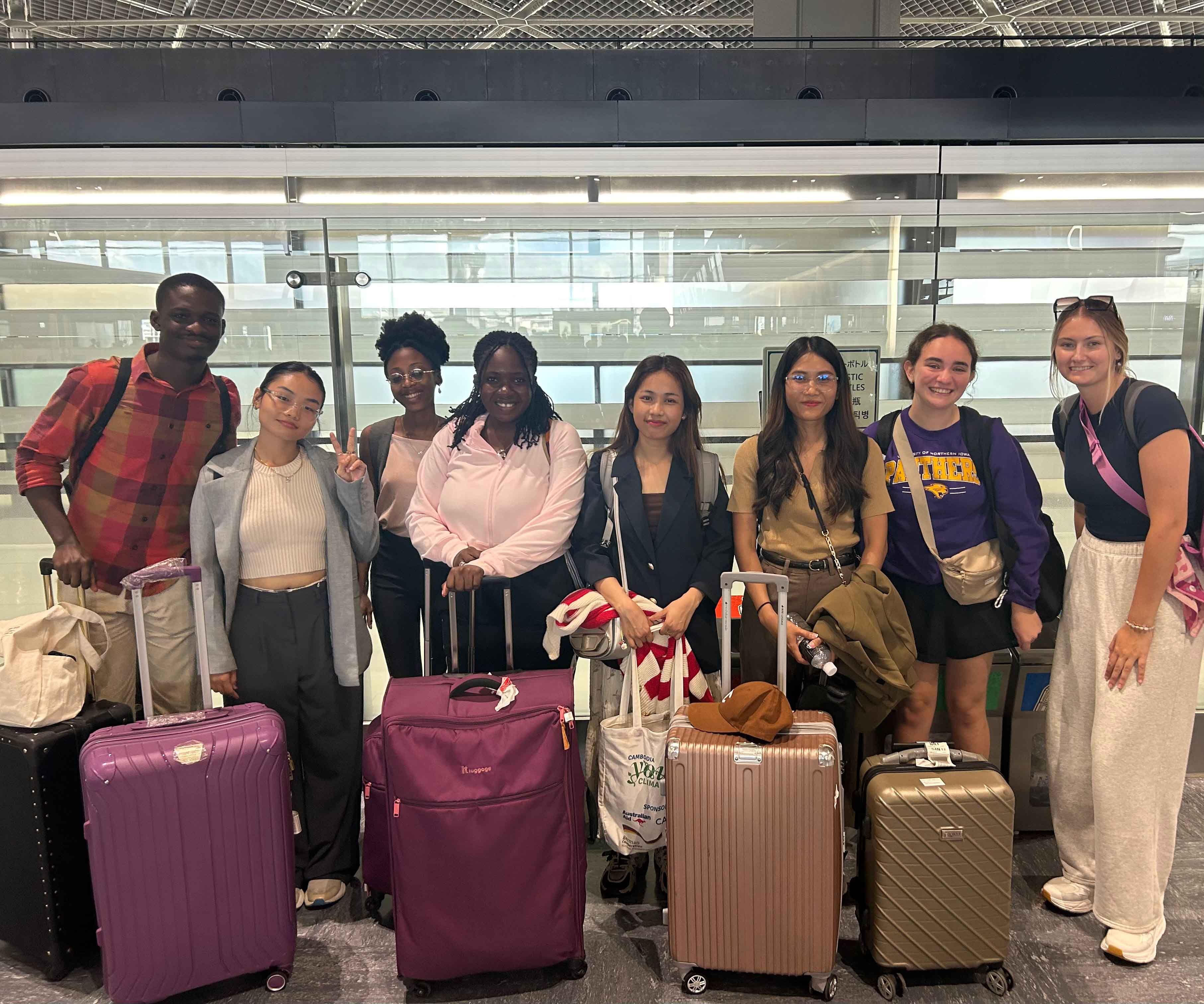A group of seven people stand together in an airport, each with a suitcase or carry-on bag. They are smiling and posing for a photo. The individuals are diverse in appearance, and several are dressed in casual travel attire. The background shows a modern airport terminal with glass walls.