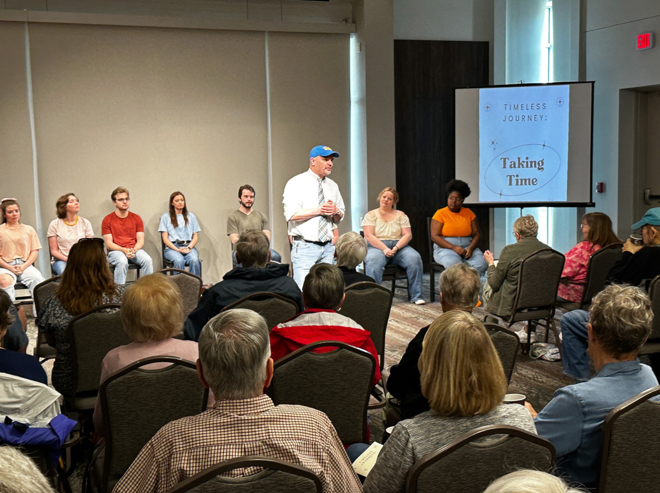 A man stands speaking to an audience in a small event room while a panel of seated participants listens behind him. A screen to the side displays a slide reading “Timeless Journey: Taking Time,” and attendees sit in rows of chairs facing the speaker.