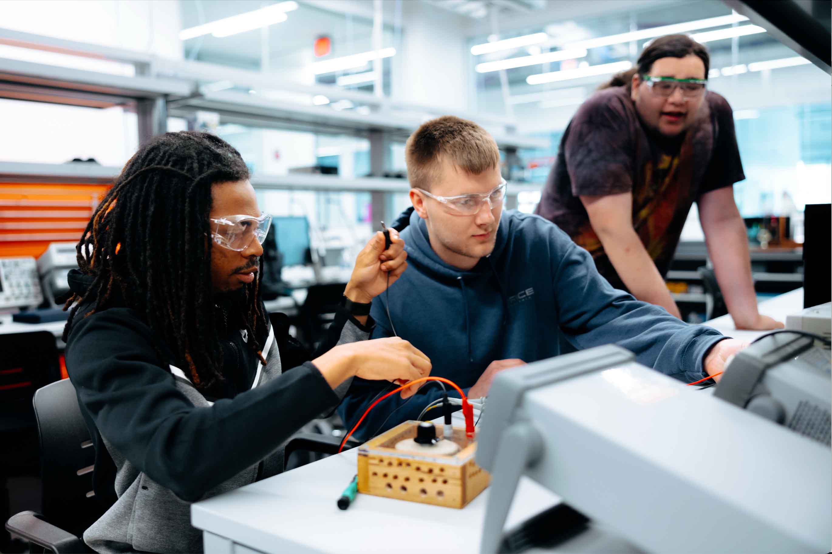 Three students wearing safety goggles work together on an electronics project in a lab. Two of them sit at a workstation connecting wires to a small wooden circuit box, while a third student stands behind them observing. The lab is filled with technical equipment and has a bright, modern setting.