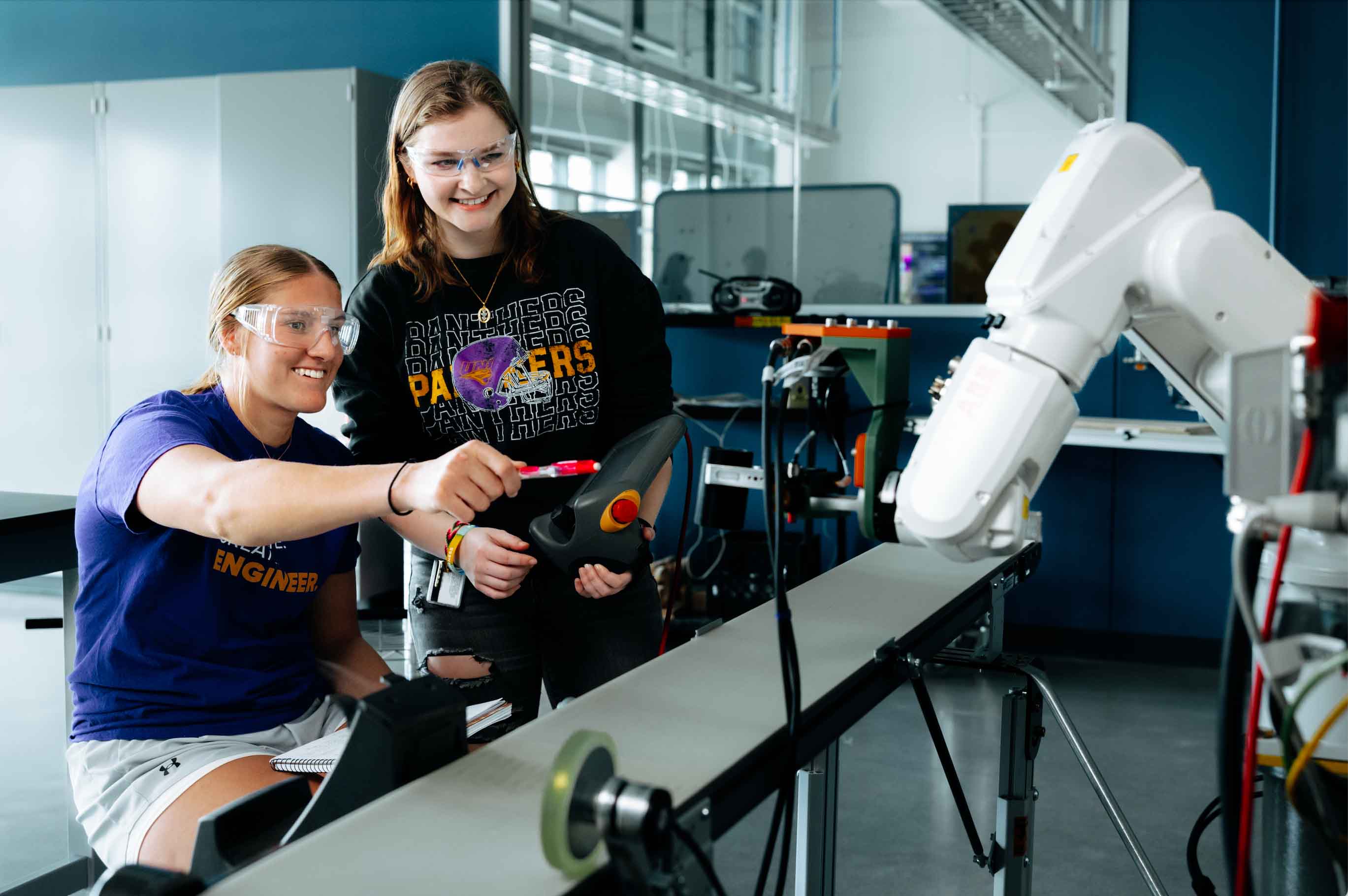 Two students wearing safety goggles work with a robotic arm in a lab. One student sits while guiding a control device, and the other stands beside her smiling and holding a controller. The robotic arm is positioned over a conveyor belt, and the background shows lab equipment and cabinets.
