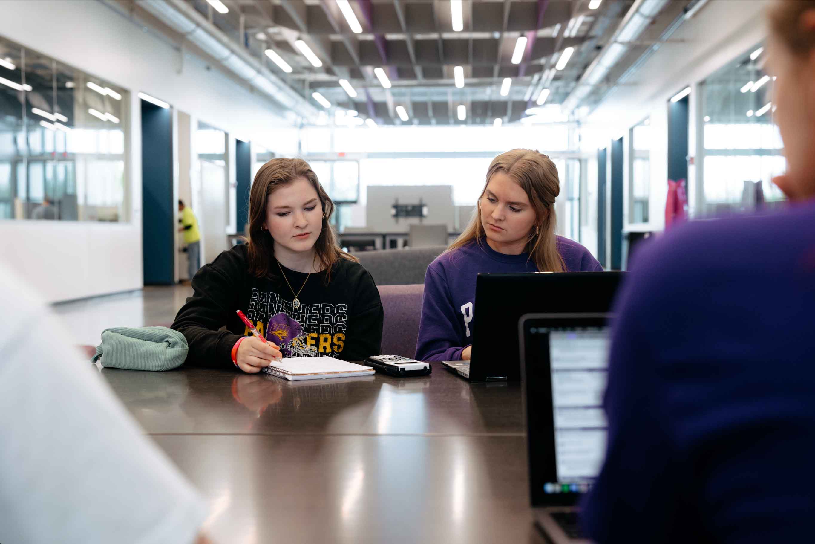Two students sit at a table in a modern, brightly lit study space. One writes in a notebook while the other works on a laptop. Both appear focused, with another person blurred in the foreground and open laptops on the table.