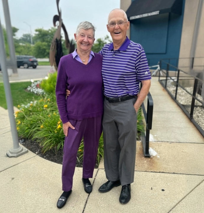 Smiling older couple standing outdoors on a sidewalk. The woman is wearing a purple sweater and matching pants, while the man is wearing a purple-striped polo shirt and gray pants. They are standing close together with flowers and greenery in the background