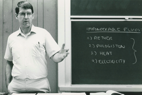 1986-11, Paul Rider teaching a Natural Science class called History & Philosophy of Science at a desk in front of a blackboard with information about imponderable fluids written on it.