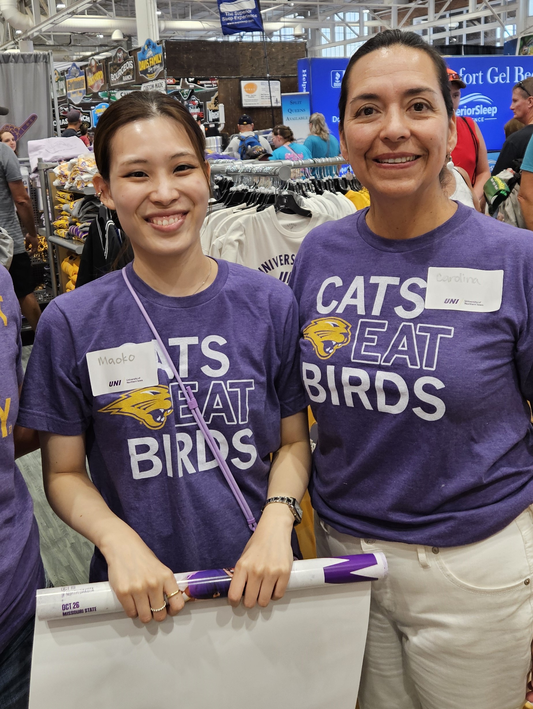 Maoko Umeda rolling a poster at the Iowa State Fair alongside Carolina Coronado-Park, both wearing Cats Eat Birds shirts