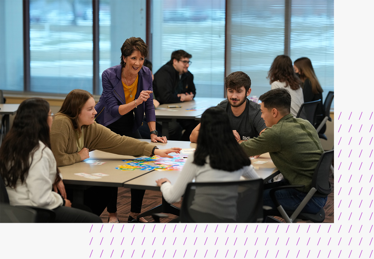 Students gather around a table during the Wilson College of Business Professional Readiness Program, engaging in a colorful card-based activity. A faculty member stands nearby offering guidance and encouragement.