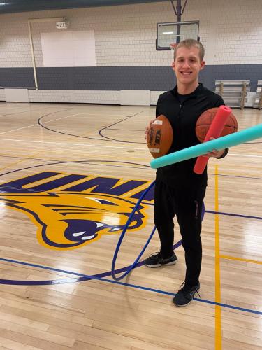 Carter Greenfield in gym carrying football, basketball and pool noodles for PE class