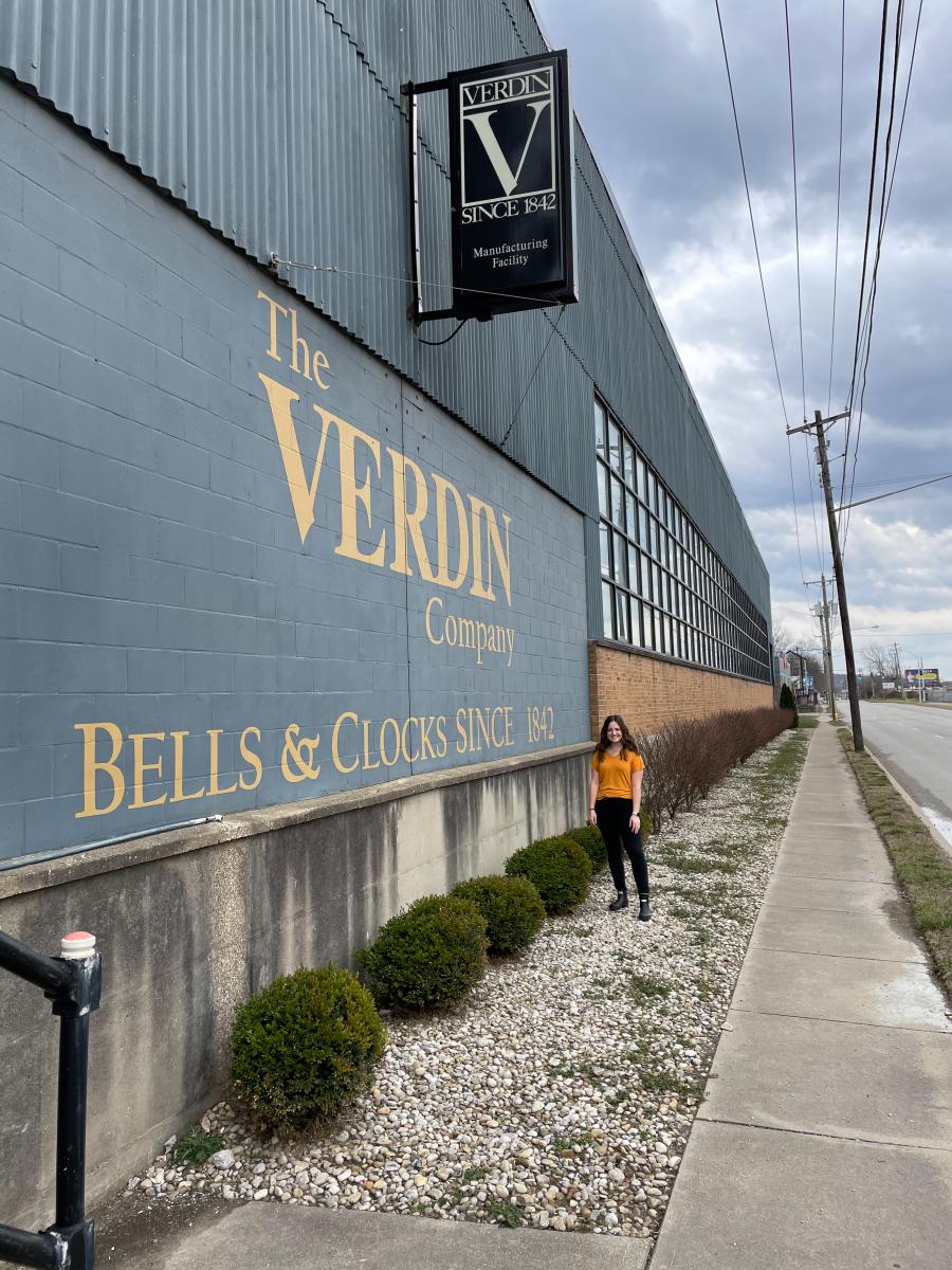 Emily Clouser in front of sign that says "The Verdin Company: Bells and Clocks Since 1842"