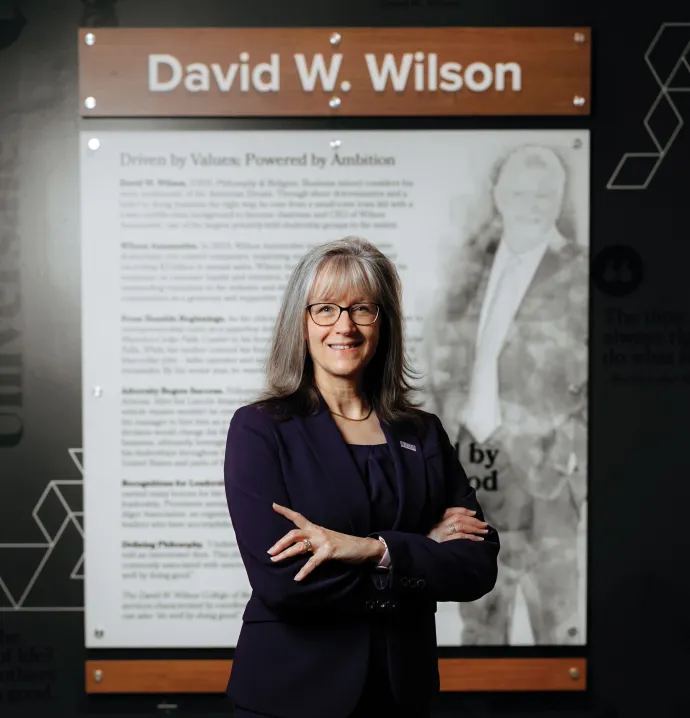 Leslie K. Wilson dean of Wilson College of business in a purple suit standing confidently with arms crossed in front of a display featuring David W. Wilson’s biography at the Wilson College of Business