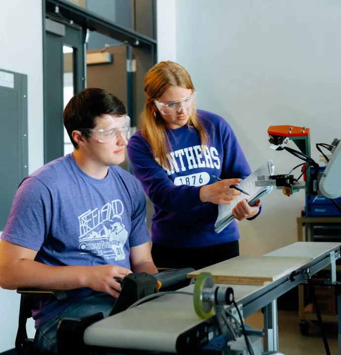 Two students wearing safety goggles work together in a lab with a robotic arm. One student sits at a control panel while the other stands beside them, pointing at notes on a clipboard. The robotic arm is positioned over a conveyor belt in a modern engineering workspace.