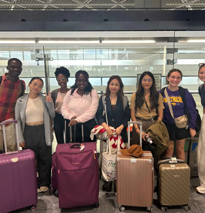 A group of seven people stand together in an airport, each with a suitcase or carry-on bag. They are smiling and posing for a photo. The individuals are diverse in appearance, and several are dressed in casual travel attire. The background shows a modern airport terminal with glass walls.