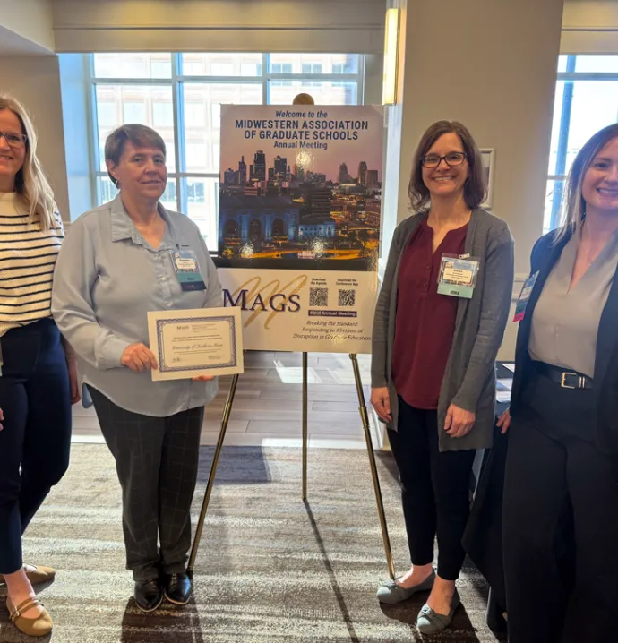 Grad Studies staff posing by MAGS signage holding Honorable Mention for the Excellence and Innovation in Graduate Education Award certificate.