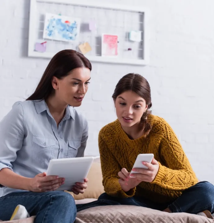 Mom on her tablet with teenage daughter by her side looking at smart phone