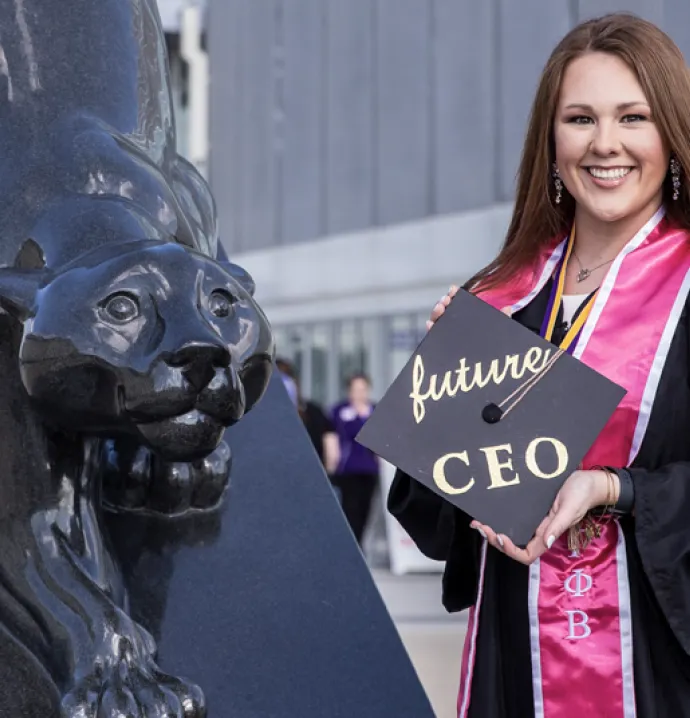 female business college student holding mortar board at commencement in front of University of Northern Iowa panther statue