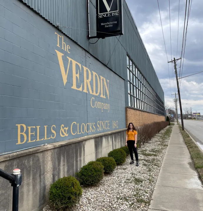 Emily Clouser in front of sign that says "The Verdin Company: Bells and Clocks Since 1842"