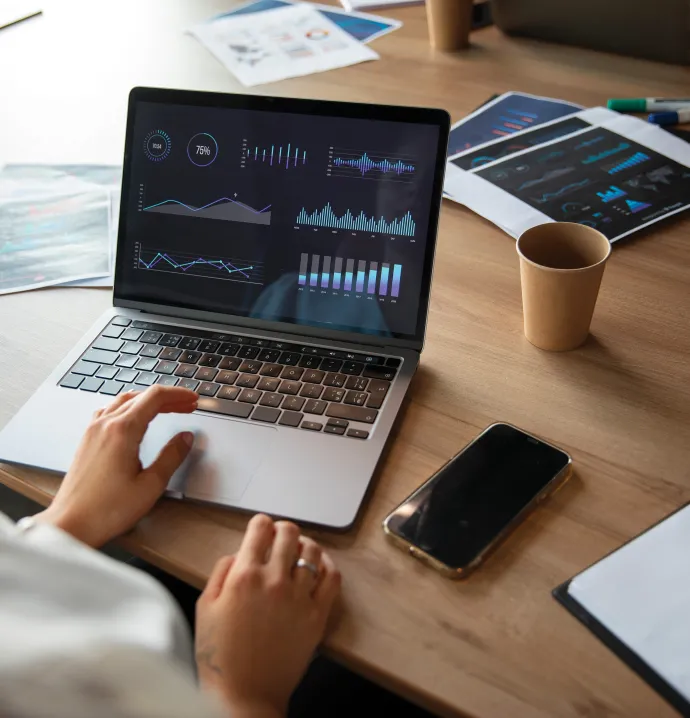 Person analyzing data visualizations on a laptop surrounded by charts and coffee cups on a desk.